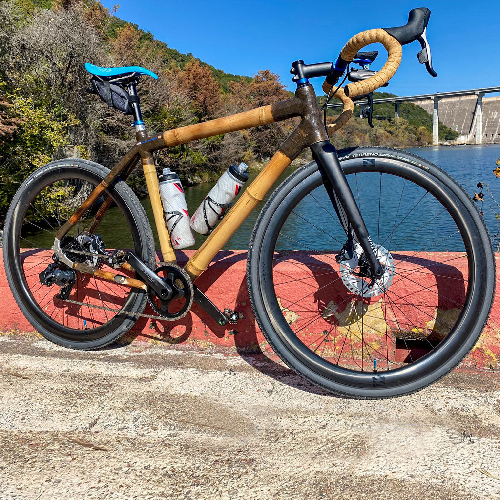 Bamboo gravel bike with bronze joints on a red parapet above a reservoir, with a dam beyond