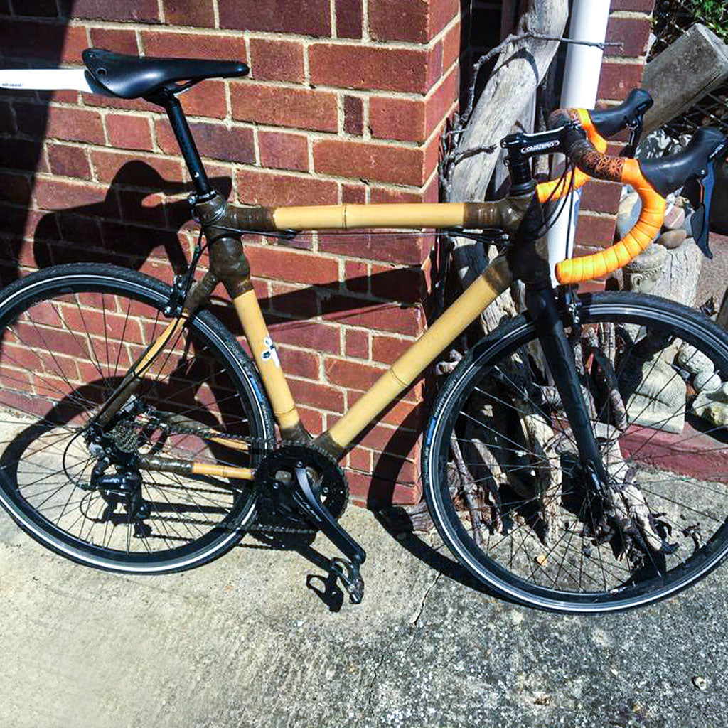 Samuel's bamboo road bike with dark wrapped joints and orange bar tape, leaning against a red brick wall