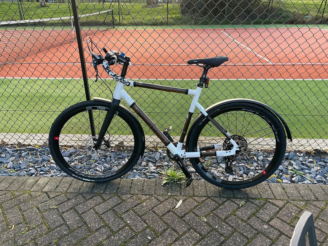 Jon's gravel bike with dark bamboo tubes, white joints and mudguards by a tennis court fence