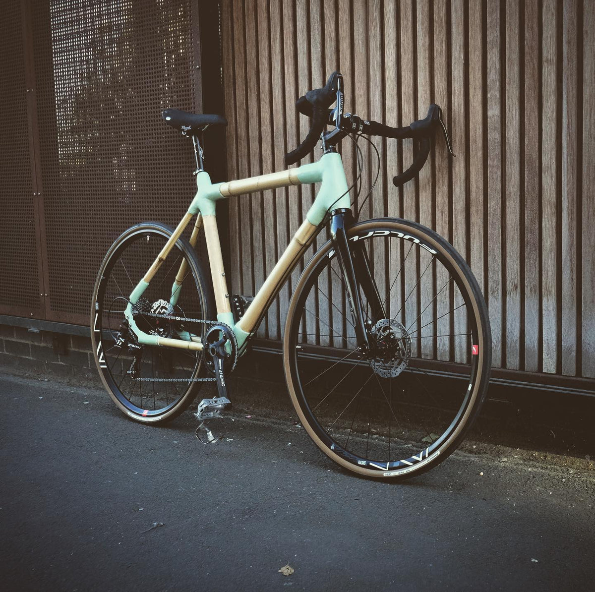 Toby's bamboo bike with mint green lugs and disc brakes, parked on a pavement by a wooden-clad wall