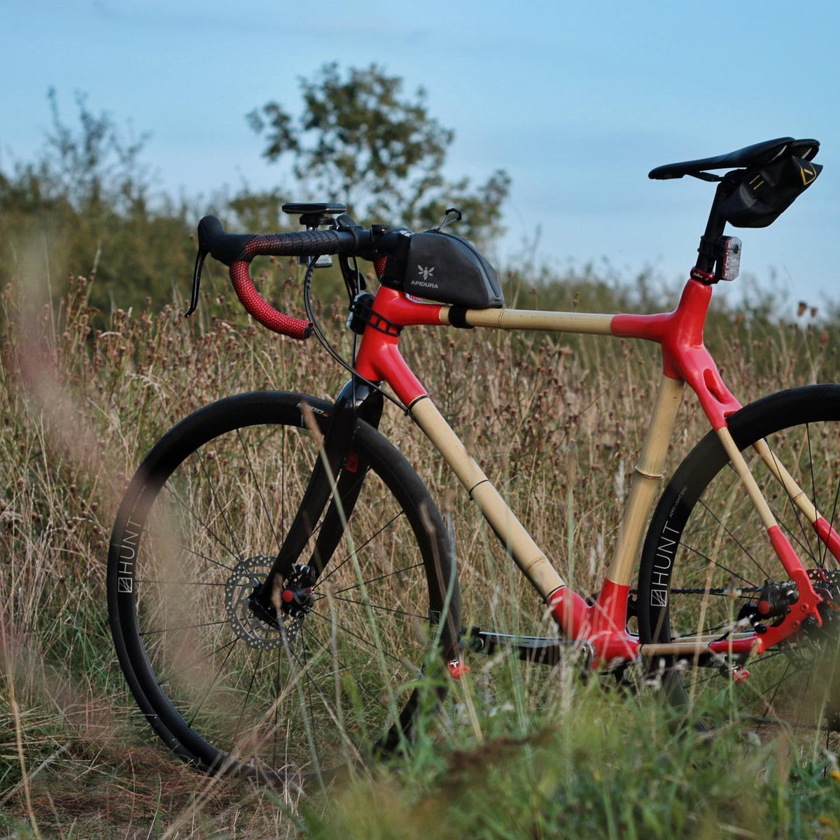 Bamboo gravel bike with red lugs, red bar tape and Hunt wheels standing in long grass