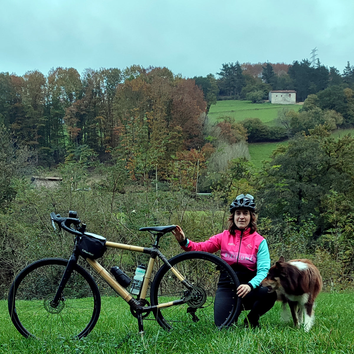 A rider and a dog beside a bamboo gravel bike on a grassy hillside with autumn trees behind