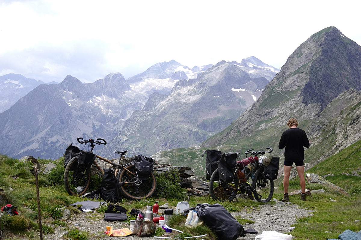 Two loaded bamboo 29ers on a mountain pass below snowy peaks, with a rider and camping kit nearby