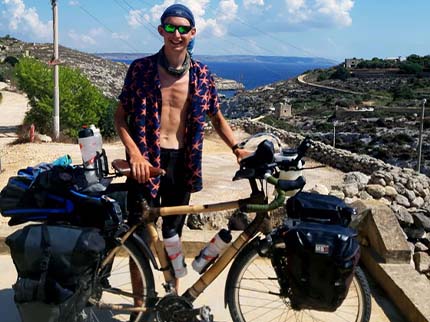 A rider standing behind Daan's bamboo touring bike, loaded with panniers, on a coastal road above the sea