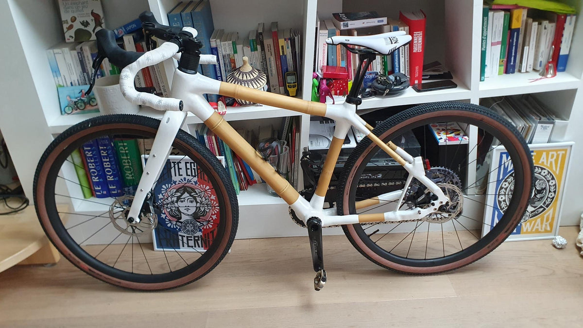 Bamboo gravel bike with white joints, white bar tape and tan-wall tyres, in front of bookshelves