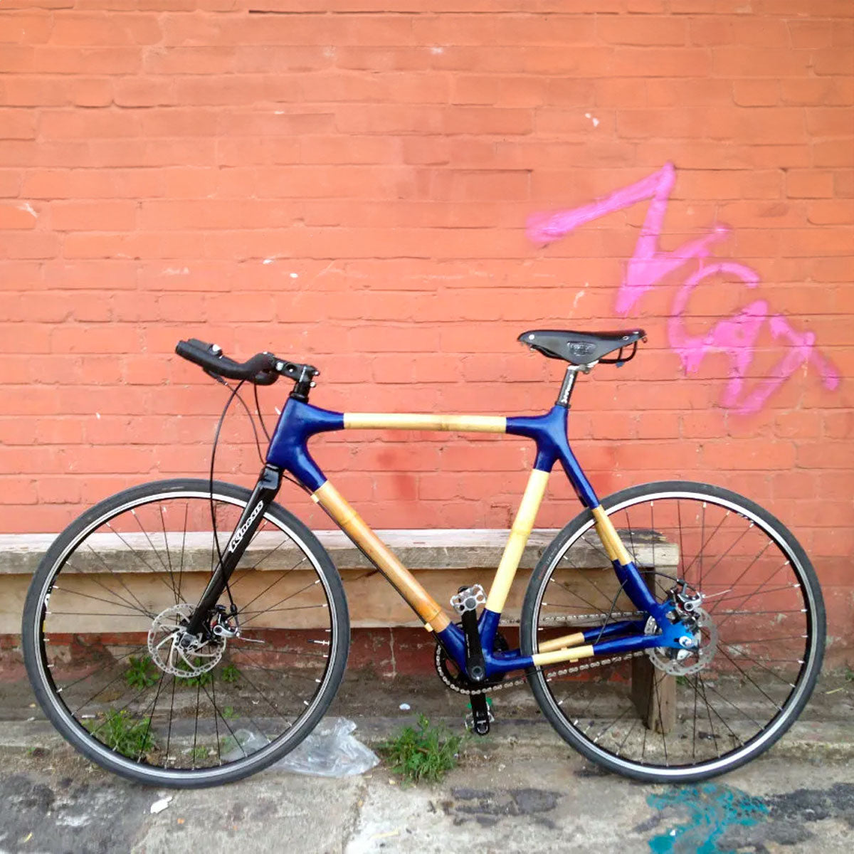Jamie's bamboo flat-bar bike with glossy blue lugs against a red brick wall with pink graffiti