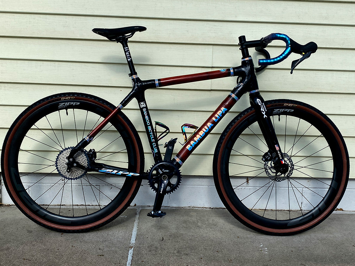 Bamboo gravel bike with a dark red finish, black joints and blue bar tape against pale clapboard siding