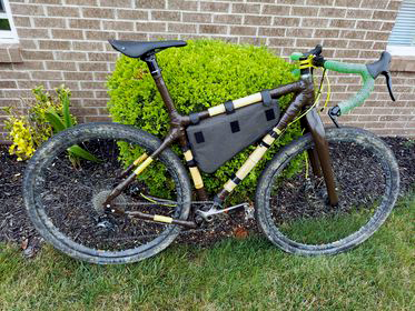 Reggie's muddy bamboo gravel bike with a frame bag and green bar tape, parked against a garden shrub