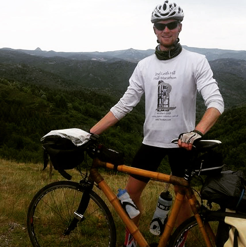 A rider stands with Tom's loaded bamboo touring bike on a hilltop above forested mountains