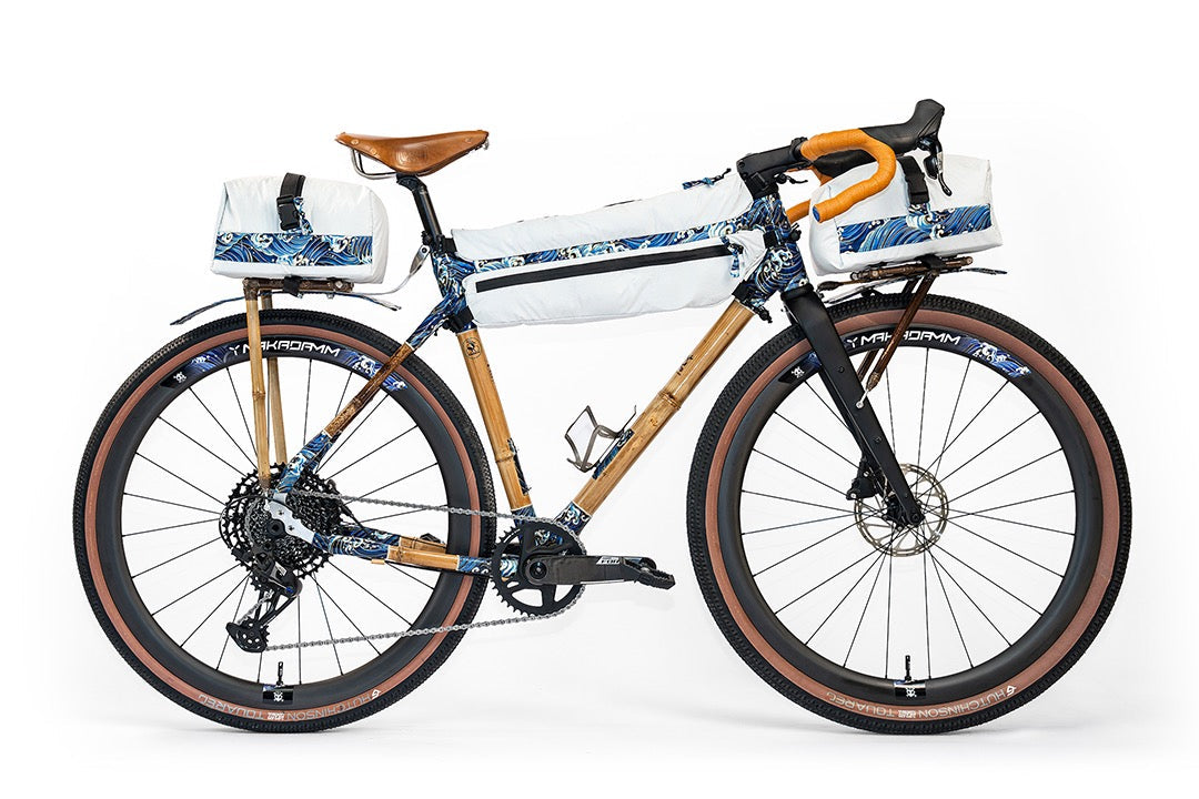 Guillaume Denneulin's bamboo bike in bikepacking mode, with knobbly tan-wall tyres, bamboo racks front and rear, and three white bags trimmed with a blue wave print