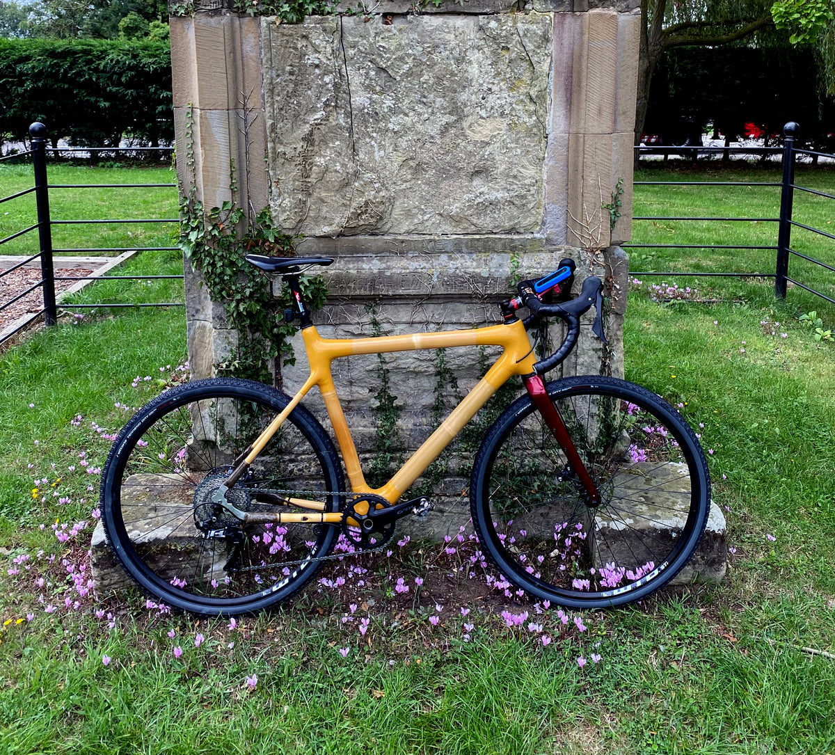 Mustard-yellow bamboo gravel bike with a red fork leaning on an old stone plinth among pink flowers