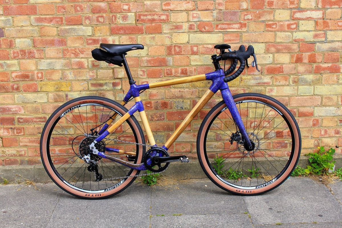 Dan's finished bamboo gravel bike, with glittery purple joints and fork, drop bars and tan-wall tyres, against a brick wall