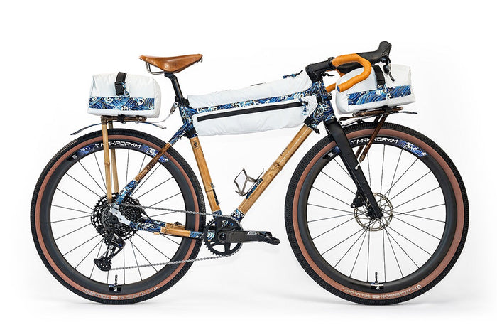 Guillaume Denneulin's bamboo bike in bikepacking mode, with knobbly tan-wall tyres, bamboo racks front and rear, and three white bags trimmed with a blue wave print