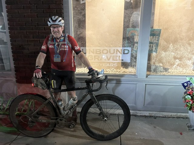 Jim Clossick at night after Unbound Gravel 200, standing with his mud-covered bamboo Gravel Lugged bike in front of an Unbound Gravel 20 years shop-window sign