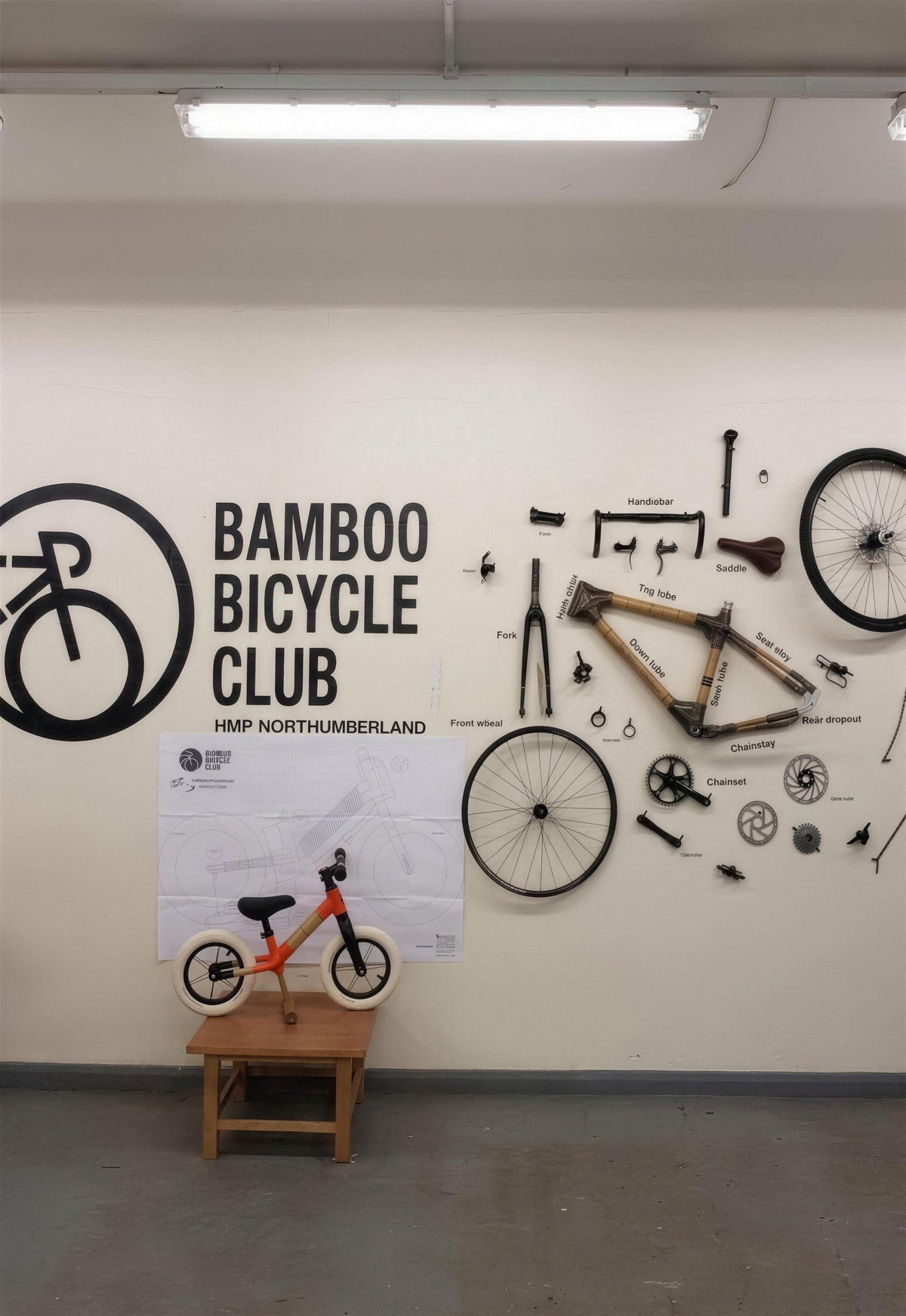 Our workshop at HMP Northumberland, the Bamboo Bicycle Club wall sign, a labelled exploded bike and a balance bike on a stool. No people.